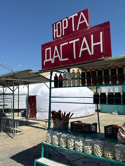 The image captures a roadside stall in a rural setting, likely in Kyrgyzstan, given the presence of yurts and the Cyrillic script on the signage. The stall displays a variety of goods, including bottles of dark liquid, likely drinks or preserved items, and jars filled with small, pale, round objects, possibly candies or traditional food items. There are also bundles of sticks in a bucket and some black crates containing jars. In the background, several white yurts are visible, suggesting a nomadic or semi-nomadic cultural context. The stall itself has a green metal frame supporting shelves. A red sign with white Cyrillic lettering, reading "ЮРТА ДАСТАН," is prominently displayed at the top, advertising the establishment. The ground is covered with gravel and dirt, and the sky is clear blue. The overall impression is that of a vibrant, authentic market stall offering local products.