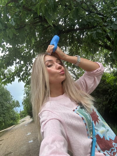 An image captures a woman with long blonde hair standing outdoors near a gravel path with puddles, likely in Armavir, Russia. She is wearing a light pink long-sleeved shirt with a colorful graphic design and a silver watch. The woman is holding a blue object to her forehead and puckering her lips, as if posing for a selfie. Overcast skies and lush green foliage suggest it might be a cloudy day. The scene appears to be in a rural or semi-rural setting, with trees and vegetation bordering the path.