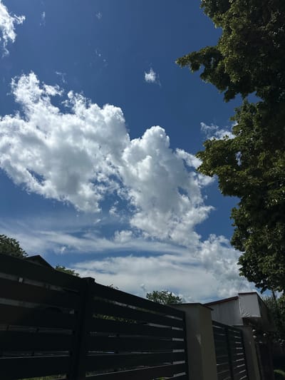 The image shows a view of the sky from a residential area in Strășeni, Moldova. The sky is a deep blue with scattered white cumulus and cirrus clouds. In the foreground, there is a dark horizontal fence made of metal slats, partially obscuring the view of a building with a white exterior and a red roof. To the right, a large tree with green foliage partially frames the scene. The overall impression is one of a sunny day in a suburban or rural setting.