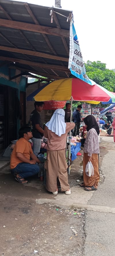 The scene takes place in a street market in Kecamatan Kutawaringin, Indonesia, located at coordinates -6.973 latitude and 107.52 longitude.  A large colorful umbrella provides shade for a stall with various items for sale. Four adults are present. One man, wearing an orange shirt and blue jeans, is crouched on the ground, seemingly talking to another man wearing a black shirt and jeans.  A woman, wearing a brown dress and white headscarf, stands near the back of the stall, facing the other man.  A woman wearing a checkered top and a brown headscarf is standing near the stall, holding a plastic bag. The stall is set up along a concrete sidewalk with a dirt and gravel road to the right. A sign with the words "DANA" and "RIK DANA" is visible on the building to the left.  Several small items, including cigarette butts, are scattered on the ground around the stall.