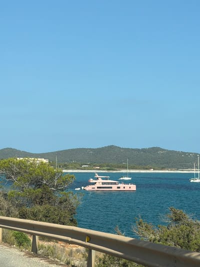The image captures a scenic view of a coastline with a tranquil blue sea under a clear sky, suggesting a bright and sunny day. The scene is dominated by a lush green, tree-covered hillside that slopes down to meet the water. The water itself is a deep blue, appearing calm and inviting.

Several boats are visible in the water, including a prominent, modern-looking pink yacht or boat with multiple decks and windows. Other smaller white boats are also present, suggesting a popular spot for maritime activities. Along the shore, there are glimpses of buildings nestled amongst the trees, indicating a coastal settlement.

In the foreground, a weathered metal guardrail runs along the edge of what appears to be a road, with dry brush and foliage growing beside it. This suggests the photo was taken from a roadside vantage point overlooking the sea. The overall impression is one of a peaceful and beautiful Mediterranean landscape, likely in a tourist destination given the presence of yachts. The time of day appears to be daylight, possibly late morning or afternoon, based on the bright lighting and clear sky. No people are visible in the image, and no text is present. The location is identified as Sant Josep de sa Talaia, Spain, which is known for its beautiful coastlines and islands.