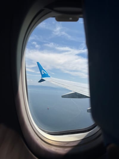 An eye-level shot through an airplane window captures a bright daytime scene from above. The dark, rounded frame of the window occupies the foreground, framing the view outside. The main subject is the white wing of an airplane, stretching diagonally across the mid-ground. Its wingtip is painted a distinct blue, featuring a white, stylized 'T' logo, characteristic of Turkish Airlines.
Beyond the wing, a vast blue sky with scattered white, wispy clouds fills the upper and left portions of the frame. Below the wing, the distant landscape or a large body of water appears hazy and light gray, suggesting considerable altitude. A notable detail in the distance, just below and to the right of the wingtip, is a small, colorful object, possibly a hot air balloon, with visible ropes or lines extending from it. Another faint, thin white line, possibly a contrail from another aircraft, is visible in the upper right sky. The overall lighting indicates a clear, sunny day.