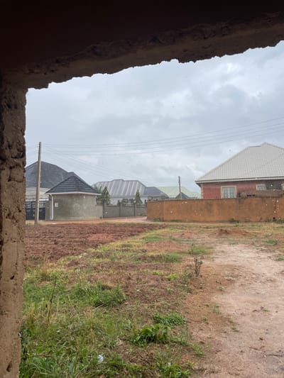 The media file presents an outdoor scene in Rafin Bauna, Nigeria, under an overcast sky. The image appears to be taken from inside or behind a rough, crumbling brown concrete or mud wall, which frames the foreground along the bottom and left edges.
In the midground, several residential buildings are visible on the left side. A prominent house features reddish-brown walls, white-framed windows, and a distinctive white patterned roof. Further down, another house, built with grey concrete, displays a dark tiled roof and a black metal gate. Electrical poles and multiple overhead power lines stretch across the mid-left portion of the frame.
To the right, a large expanse of unpaved, reddish-brown earth mixed with sparse green grass and weeds extends into the background, suggesting an undeveloped plot of land or field.
The sky is uniformly light grey and heavily clouded, indicative of an overcast day. Numerous small, white, circular specks are scattered across the entire image, most noticeably against the sky and darker surfaces, suggesting light precipitation such as rain or sleet. There are no people visible, and no discernible text appears in the scene. The overall impression is one of a quiet, somewhat damp, daytime view of a developing residential area.