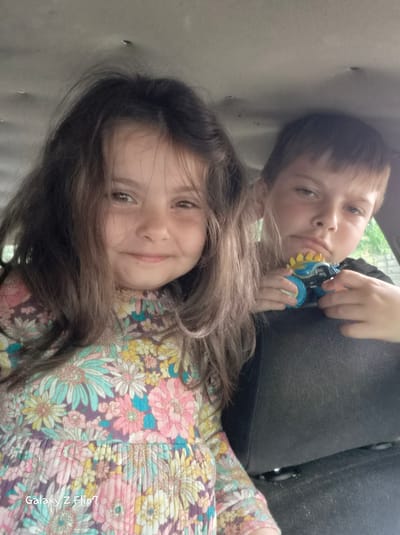 The image, rotated 90 degrees clockwise, features two children, a girl and a boy, appearing to be in an enclosed space, possibly a car interior. The setting is Kalipetrovo, Bulgaria.

The girl is positioned at the top of the frame (left in the original orientation) and has long, dark hair. She is wearing a colorful floral dress with patterns of pink, light blue, yellow, and white flowers on a purple background. She is looking directly at the camera with a clear smile, showing a cheerful emotion.

Below her is a boy with short, light brown hair, wearing a dark top. He is holding a distinctive blue and yellow toy with gear-like or spiked features. The boy also looks towards the camera, but with a more neutral expression. Behind him, through what appears to be a window, a hint of green foliage is visible, suggesting an outdoor environment beyond the immediate space.

The background immediately surrounding the children is a plain, light-colored surface, possibly a car ceiling or headliner, with some minor dark blemishes. Dark fabric, likely part of a car seat or interior, is also visible. The lighting is soft and even, indicating it's likely daytime, perhaps indoors or on an overcast day. The visible text "Galaxy Z Flip7" is a watermark on the girl's dress, indicating the camera used. The children appear to be posing for the photograph.
