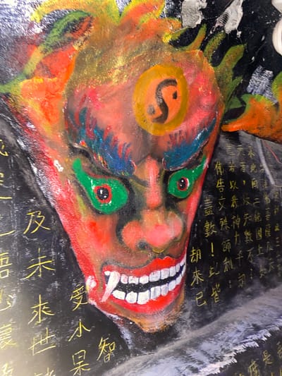 The image shows a close-up of a colorful, painted mask or mural depicting a fierce, demonic face. The face is predominantly red with bright green eyes, sharp white teeth, and fiery orange and yellow hair or headdress. There are Chinese characters painted in gold on the black background surrounding the face. The mask appears to be mounted on a textured surface, possibly a wall or a structure. The image's metadata indicates it was taken in Xuan Thanh City, Vietnam, suggesting this is a cultural or religious artwork found in that location.