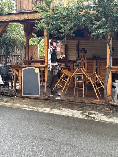 A mannequin dressed as a cowboy stands on a wooden porch of a rustic building. The cowboy mannequin wears a brown cowboy hat, a red bandana, a white shirt with a floral print, a dark vest, and brown pants. A leather pouch is attached to its waist. In front of the mannequin is a chalkboard sign with a yellow "Glovo" sticker. To the left of the mannequin, a black barbecue grill with a metal chimney stands on a cart with wheels. Several wooden stools are placed haphazardly on the porch, some stacked on top of each other. The building behind the porch has wooden siding and an overhead structure with wooden beams. Trees with green leaves overhang the scene. The ground in front of the porch is paved with asphalt, which appears wet, suggesting it may have rained. The overall scene depicts a western-themed establishment, possibly a restaurant or bar. The location is identified as Sveti Vlas, Bulgaria.