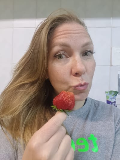A blonde woman with green eyes and a grey t-shirt is holding a strawberry to her lips. She has long, wavy blonde hair that falls over her shoulder. The background is a white tiled wall. There is a green logo on her t-shirt that appears to say "boost". The image is a selfie, and the woman is looking directly at the camera. The location is in the city of Kemul, Russia.