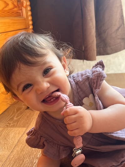 This eye-level, close-up image, set in Elena, Bulgaria, features a happy young child, likely a baby or toddler, looking directly into the camera. The child has fair skin, wide, dark eyes, and wispy brown hair. They are wearing a light purple or mauve-colored dress or romper, made from a textured, possibly crinkled, fabric with ruffled edges around the shoulders and waist. A subtle white daisy-like flower design with a yellow center is visible on the side of their garment.

The child is actively holding and chewing on a pink pacifier clip. The clip features a small, embossed cat-like face design on its plastic component, attached to a metal clasp. The ribbed, teether portion of the clip is partially inside the child's mouth, which is open in a playful expression, revealing their tongue. Their overall demeanor suggests happiness and engagement.

The background indicates an indoor home setting. A brown corduroy-textured sofa or piece of furniture is visible in the upper left portion of the image. The flooring is a prominent feature, consisting of light-colored wooden laminate or parquet with a visible grain pattern, extending across much of the background and foreground. A segment of wooden furniture or detailing with a scalloped or decorative edge can also be seen in the bottom left corner. The lighting appears bright and natural, consistent with daytime. No visible text is present in the image.