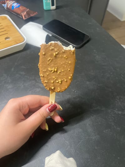 A person's hand with red and yellow nail polish holds a bitten chocolate-covered ice cream bar with nuts. The ice cream bar is on a wooden stick. The background is a dark, speckled countertop. In the background, out of focus, are a white object, a dark smartphone, and a small teal and white device. Also visible is a wrapper from another chocolate bar. The image is cropped to show the hand and ice cream bar prominently, with the background elements less clear. The scene appears to be indoors, likely in a domestic setting.