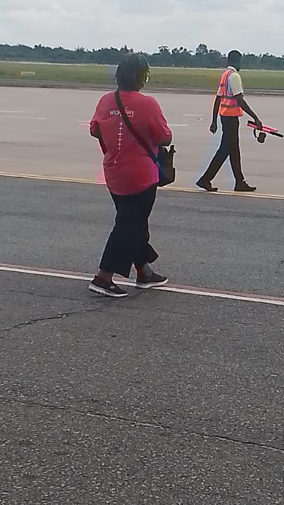 The image shows two individuals walking on an airport tarmac. The person in the foreground is wearing a bright pink t-shirt with a graphic design on the back that includes the text "WEST AIR" and several small airplane icons. They are also wearing black pants and dark sneakers. A dark bag is slung over their shoulder. The second person, further in the background, is wearing a high-visibility orange vest over a light-colored shirt and dark pants. They are carrying a pink object with a black handle, possibly a signaling device or tool. The background features an open expanse of tarmac, with a line of trees visible in the distance under a cloudy sky. The scene appears to be outdoors during the daytime, likely at an airport given the setting and the presence of airport personnel. The overall mood is neutral, depicting everyday activity at an airport.