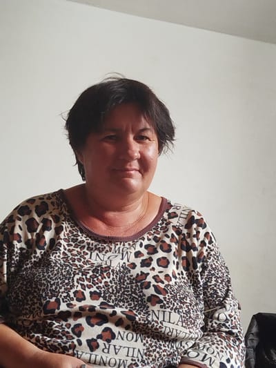 The media file is a medium shot, horizontal photograph of a middle-aged woman posing against a plain, light-colored wall. She has short, dark hair, fair skin, and is looking towards the camera with a slight smile. The woman is wearing a long-sleeved shirt with a leopard print pattern and a repeating text print on it. The text appears to be a repeating word or phrase, but it is not clearly legible due to the print and resolution of the image. The lighting in the image suggests it was taken indoors during the daytime, likely with natural light. There are no other objects or people visible in the frame, and the background is nondescript, offering no specific location cues beyond it being an interior space. The woman's expression is calm and relaxed, indicating a posed, informal portrait. The overall setting is simple and domestic. The location context provided is "Зиянчурино, Russia".