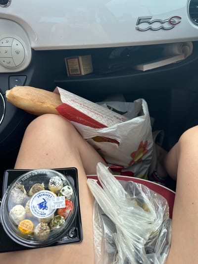The image is a close-up, eye-level shot taken inside a Fiat 500 car, likely in Ouville-la-Rivière, France, given the text on the baguette packaging. In the foreground, a person's bare legs are visible, positioned as if sitting in the driver's or passenger's seat. A black plastic container filled with small, assorted cheeses is placed on the person's lap. Next to the cheese, a clear plastic bag contains what appear to be green olives. Behind the person, a white plastic shopping bag with a red and yellow flower design holds a baguette, which protrudes from a red and white paper wrapper. The car's interior is visible, including the dashboard with the "500" logo, climate control buttons, and a glove compartment containing a small box and a rolled-up document. The scene suggests a casual stop, possibly for a picnic or a snack, with groceries purchased from a local market or boulangerie.