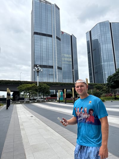 A man in a blue t-shirt and patterned shorts stands on a paved walkway in front of several modern, glass-facade skyscrapers. He holds a smartphone in his right hand. The t-shirt features a graphic of several people and the text "Ready Play Australian Open 27 January 2019." In the background, a multi-level bridge covered in greenery spans across the scene, with trees visible beneath it. The sky is overcast. The architecture suggests a contemporary urban environment, consistent with modern developments in the Huangpu District of China.