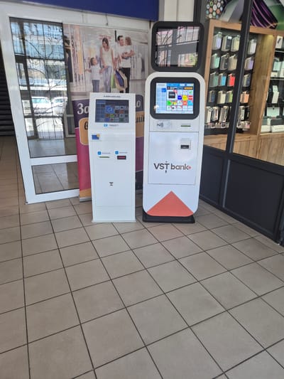 This media file shows two white payment kiosks positioned in front of a store display filled with various mobile phones. The kiosk on the left has a touch screen displaying a grid of colorful icons for different applications and services, along with a phone number "0 800 600 350" and a product number "ND 1065221". The kiosk on the right features the VST bank logo, a card insertion slot, and a receipt dispenser. Both kiosks have a contactless payment symbol.
Behind the kiosks, a large advertisement featuring a smiling family with shopping bags is visible. The floor is tiled with a grid pattern. The setting appears to be a commercial space, possibly inside a shopping mall or a store, given the display of electronics and the overall layout. The lighting suggests it could be daytime. The presence of the VST bank logo indicates a financial service point. The overall scene conveys a sense of modern retail and accessible banking services. There is also text in Cyrillic script visible on the kiosk and the advertisement.