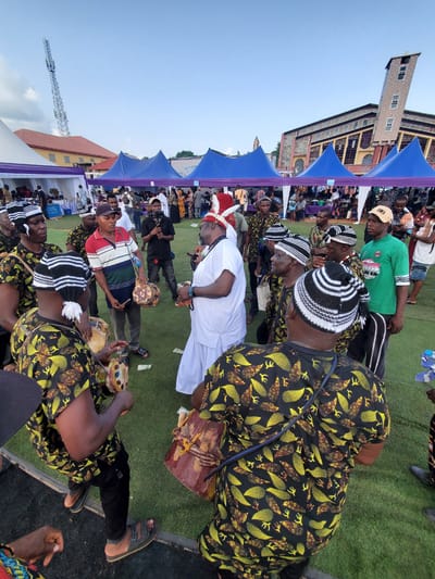 A low-angle, wide shot captures a vibrant outdoor gathering in Awkuzu, Nigeria. Predominantly men fill the frame, several positioned in the foreground. Many wear distinctive black-and-white striped hats and patterned shirts. Some are playing drums, and one man, clad in white, stands out with a red-feathered headdress. Other individuals stand in the background, some of whom are filming. Several blue tents stand along the left side of the frame, with a multi-story brick building in the background. The setting includes an area covered in green turf, and a clear, slightly overcast sky completes the composition.