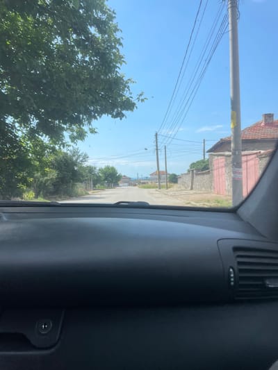 The media file captures a view from inside a vehicle, looking out towards a street scene in Malevo, Bulgaria. The camera is oriented vertically, making it appear as if the viewer is looking down a road from a high vantage point. The sky is a clear, bright blue, suggesting a sunny day.
On the left side of the frame, lush green trees with dense foliage are prominent. The road itself is paved and appears to be a residential street, lined with houses and utility poles. Several power lines are strung between the poles, adding to the rural or suburban feel of the location. Some of the houses have red-tiled roofs and stone or light-colored walls, with some featuring closed, red-colored garage doors. The architecture suggests a modest, possibly older, settlement.
In the foreground, on the right side of the image, the interior of the car is visible. This includes a dark gray dashboard with a ventilation grille and what appears to be a control knob or button. The edge of the car's window frame is also present, indicating the perspective is from a passenger or driver's seat.
There are no people clearly visible in the scene. The overall impression is that of a peaceful, everyday scene during daylight hours in a Bulgarian village or town. The image does not contain any discernible text.
