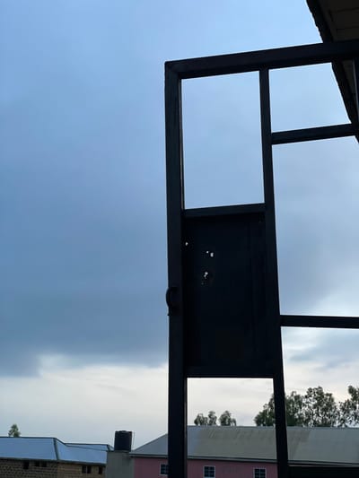 The image shows a dark, rectangular metal frame against a cloudy sky. The frame appears to be part of a larger structure and has a door-like opening. On the left side of the frame, there are several small, irregular holes. In the background, several buildings with corrugated roofs are visible, as well as a water tank. Trees line the horizon behind the buildings. The scene is consistent with an outdoor setting in Wumat, Nigeria.