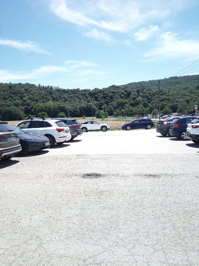 The image depicts a parking lot in Besalú, Spain, with a dense, green forest covering a hillside in the background. Several cars are parked in rows on a light-colored, gravelly surface. The sky is a bright blue with scattered white clouds. The foreground is dominated by the textured surface of the parking lot.