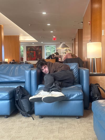 The image captures an indoor scene, likely a lounge or common area, with a relaxed atmosphere. The perspective is from a slightly elevated position, looking across a row of modular seating.
In the foreground, a young person with dark, shaggy hair and light skin is seen lying face-down across two blue, upholstered lounge chairs. They are wearing a dark brown hooded sweatshirt, dark pants, and light-colored sneakers with dark soles. Their body is bent at the waist, with their legs extending upwards, and their head is turned towards the viewer, revealing a calm, perhaps slightly amused, expression. One black backpack is visible on the carpeted floor next to the chairs, near the person's feet.
The room features a combination of light-colored walls and warm wooden paneling. The floor is covered with a light brown patterned carpet. Overhead, several recessed lights illuminate the space, and a tall floor lamp with a white rectangular shade stands near the wooden wall paneling.
In the midground and background, more blue lounge chairs are arranged, some occupied by other individuals who are mostly indistinct but appear to be seated or lounging. Through large windows at the back, green foliage from trees is visible, indicating an outdoor view and suggesting it is daytime. A large, abstract painting with rich red, blue, and dark tones hangs on the wall below the windows. Further in the distance, a red "EXIT" sign is visible on a white wall. Architectural details like triangular-shaped elements or light fixtures can also be seen higher up in the background. The overall impression is one of a comfortable and casual public space.