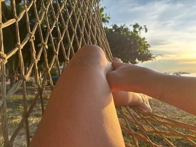The image depicts a first-person perspective from an individual resting in a light brown woven net hammock in Koh Tao, Thailand, facing towards the sea during either sunrise or sunset. The foreground features the individual's bare legs and right arm. The left leg is bent, displaying a faint, faded linear tattoo on the upper thigh. The right forearm, resting across the left leg, exhibits a delicate, branching tattoo resembling a tree or nerve structure. The hammock's knotted cord netting, forming diamond patterns, is prominent on the left and bottom. Beyond the hammock, a grassy area with palm trees and tropical foliage is visible. The background shows the sea with multiple vessels silhouetted against a brilliant golden sky, characteristic of low sun angle. No other individuals are present.