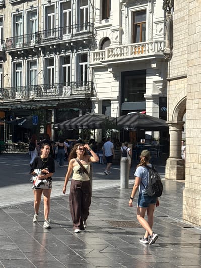 This image captures a street scene in Brussels, Belgium, likely on a sunny day. In the foreground, three women are walking on a paved square. The woman on the left is wearing a black t-shirt, striped shorts, and sneakers, and is holding some papers. The woman in the middle is wearing a light-colored strapless top, loose brown pants, and sandals, and has her hand on her hair. The woman on the right is wearing a light blue t-shirt, denim shorts, a backpack, and athletic shoes, and is walking away from the camera.
In the background, there are buildings with architectural details typical of European cities, including balconies and ornate windows. A row of outdoor seating with umbrellas suggests a cafe or restaurant. Other pedestrians and part of a stone building with an arched opening are also visible, contributing to the urban atmosphere. The lighting suggests it is midday.