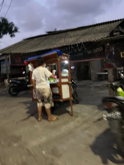 A person stands behind a food cart at dusk in Kludan, Indonesia. The person is wearing a white t-shirt and patterned shorts, and is reaching towards the cart. The cart is wooden with a blue tarp and appears to be selling food. A sign on the building behind the cart reads "PRIMA 16.000". Motorcycles are parked on the street. The sky is overcast and the lighting suggests it is either early morning or late evening.