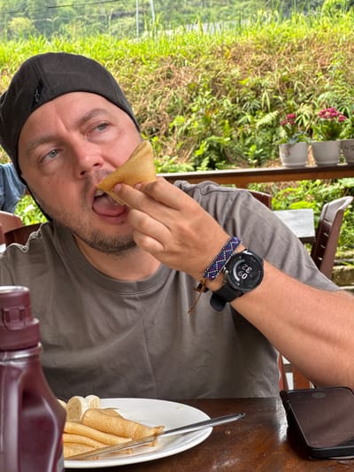 An outdoor, eye-level shot in Du Già Commune, Vietnam, captures a man in a grey t-shirt and dark grey cap eating what appear to be pancakes or crepes. He is bringing a folded piece of food to his open mouth with his right hand. On his left wrist, he wears a black smartwatch displaying "0:30" (likely 12:30 PM given the bright daylight), "99%" battery, "72" heart rate, and "113" steps, along with "TUE" for Tuesday. Below the watch is a woven bracelet with purple, blue, and white patterns.

The man's cap has a small white star-like logo on the front. He has light eyes and short facial hair. To his right, a black smartphone rests on a wooden table. A white plate on the table holds more folded pancakes/crepes and several slices of banana, with a spoon or fork on the plate. In the foreground, a brown squeeze bottle, possibly containing syrup or sauce, is visible. The background features lush, bright green foliage, indicating a natural, outdoor setting, possibly a cafe or eatery overlooking greenery. The man appears focused on his meal.