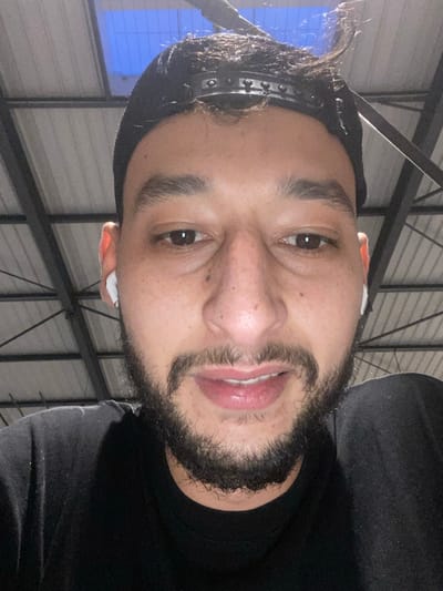A man with a beard, dark hair, and dark eyes faces the camera. He wears a black beanie with a silver-studded band and has a white wireless earbud in his right ear. He wears a black shirt, and the background features an industrial ceiling with exposed metal beams and a window with blue and white light. The image is vertically oriented.