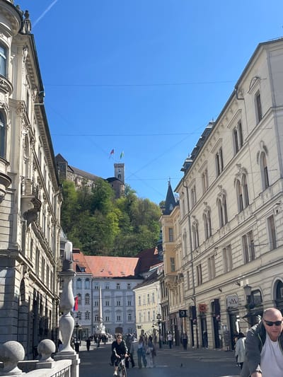 The image shows a street in Ljubljana, Slovenia, looking up towards Ljubljana Castle. The street is lined with buildings with ornate facades. The castle is perched on a hill overlooking the city, with flags visible on the tower. People are walking and cycling on the street. The sky is clear and blue.
