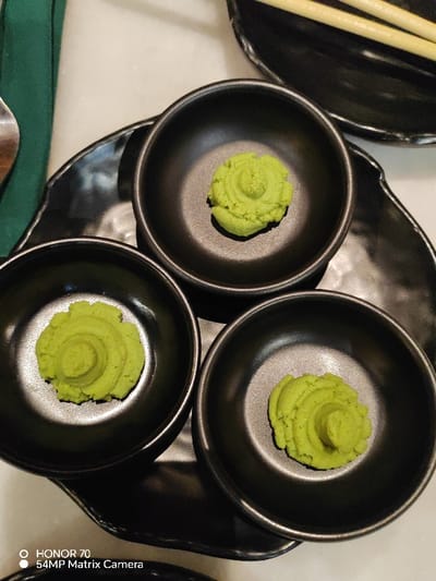 Three small, black bowls are arranged on a larger, decorative black dish. Each bowl contains a vibrant green dollop of wasabi. The wasabi is textured with a ridged pattern, resembling a small mound. In the background, a set of chopsticks rests on another black dish. A green napkin and a spoon are partially visible on the side. The surface beneath is a light cream color.