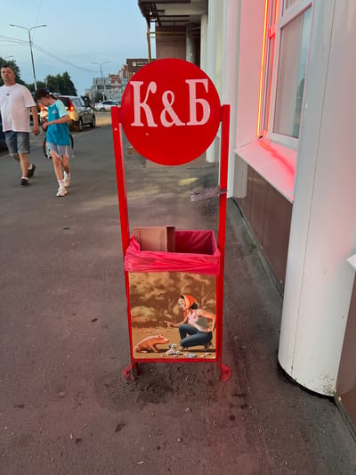 The image depicts a street scene in Cheboksary, Russia, with a prominent red trash receptacle in the foreground. The receptacle features a large red circular sign with white Cyrillic letters "K&B" and a colorful illustration at its base. The illustration shows a woman in a pink tank top and blue jeans, wearing an orange headscarf, crouching down to interact with a piglet and scattered items on the ground.
Two individuals, an adult male and a child, are walking on the asphalt sidewalk to the left of the trash receptacle. The man wears a white t-shirt and denim shorts, while the child is dressed in a blue and turquoise t-shirt and denim shorts. Behind them, several cars are visible on the street, and buildings line the background. The lighting suggests it is either early morning or late evening. The overall environment appears to be an urban setting.