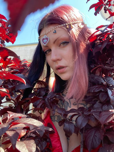 An upward-angled, close-up portrait features a person with pink/rose gold hair, framed by dense, dark red foliage and a bright blue sky. The individual, looking directly at the camera with a neutral expression, wears a decorative gold chain headpiece adorned with small charms such as stars, a sun, and black hearts. A prominent red heart-shaped medallion featuring an "evil eye" design is visible near their temple.
Their eyes are light-colored, enhanced with dark eyeliner and mascara. Partially visible tattoos can be seen on their neck and shoulder, intertwined with the dark red leaves. A piece of red fabric or clothing is visible on the person's right side. The foliage, possibly from a red-leafed tree or bush, is brightly illuminated by direct sunlight, creating highlights and shadows and partially obscuring the figure. The background is a clear, light blue sky. The strong, direct light indicates a daytime setting, likely midday or early afternoon, under sunny conditions. The scene is set outdoors in Kunje, Montenegro. No visible text is present in the image.