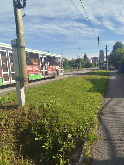 A bright, sunny day in Zavodoukovsk, Russia, features a modern, white and green public bus with Cyrillic advertisements visible on its side. The bus is traveling on a paved road next to a grassy area and sidewalk. A wooden utility pole with attached wires and a small security camera stands prominently in the foreground. In the background, trees, buildings, and a distinctive industrial structure contribute to the urban landscape under a blue sky with wispy clouds. The scene captures a typical moment of urban transportation and infrastructure within the city.