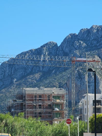 In Deveses, Spain, a construction scene unfolds against the backdrop of a rugged mountain range. A multi-story building, covered in scaffolding and constructed with red brick, is in the foreground. Adjacent to it, a modern white building is also under construction, with visible scaffolding. A large tower crane, with the logo "CANEVERAL CRANES" visible on its arm, dominates the skyline. A red stop sign is positioned in front of the buildings. Lush green foliage lines the bottom of the frame, suggesting a transition from wild vegetation to a developed area. The sky is clear and blue.