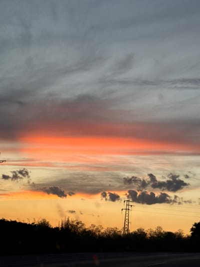 The image captures a sunset over a landscape, likely in or near Veselina, Bulgaria. The sky is a dramatic display of color, with a band of bright orange clouds stretching horizontally across the frame. Above this fiery band, the clouds transition to a gradient of grays, suggesting an approaching storm or the lingering darkness of twilight. Below the orange glow, the sky is a pale yellow, fading into the dark silhouette of trees and vegetation that form the horizon line. A prominent power line tower stands tall against this backdrop, its lattice structure clearly visible. The foreground shows a dark road, hinting that the photograph was taken from a moving vehicle. The overall scene evokes a sense of natural beauty and the transition from day to night.