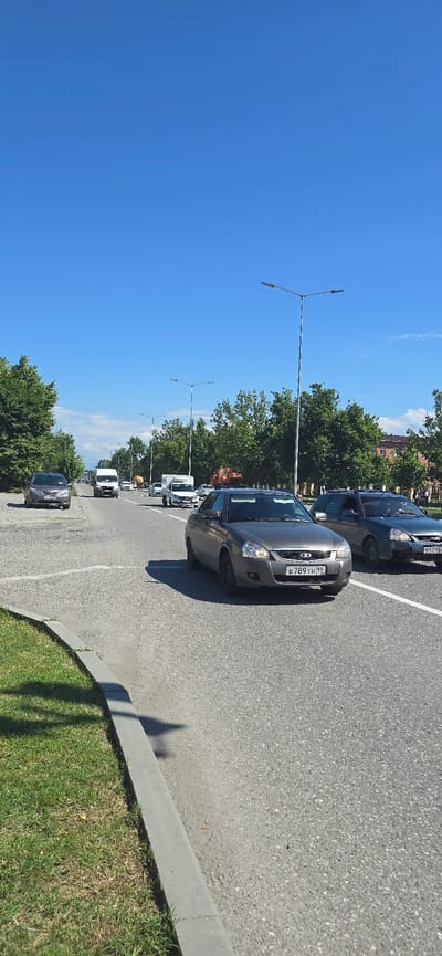 The image shows a street scene in Grozny, Russia, under a clear blue sky with sparse white clouds. A multi-lane asphalt road runs through the center, bordered by a grass verge and a curb on the left. Tall trees line both sides of the road, with some buildings visible in the distance on the right.

Several vehicles are present on the road. In the foreground on the right, a dark-colored car is turning right. In the center foreground, a dark gray sedan with license plate B 789 TH 95 is driving forward. To its right, a dark green station wagon is also driving forward. Further down the road, several white vans and other cars are visible, suggesting moderate traffic. A large orange construction vehicle is parked on the right side of the road in the mid-ground. Streetlights are positioned at intervals along the road. The overall impression is a sunny day in an urban or suburban setting with active road traffic.