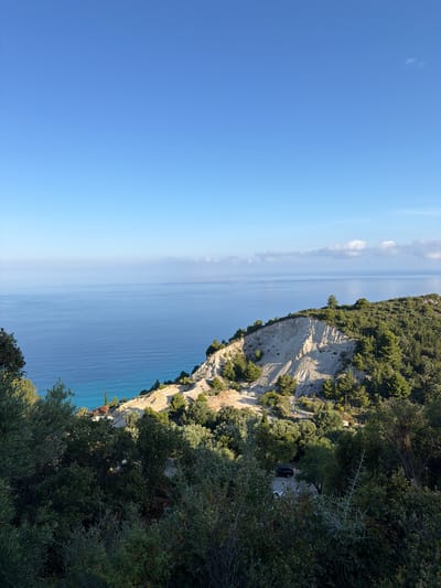 The image shows a coastal scene in the Municipal Unit of Sfakiotes, Greece. A cliff face covered with vegetation drops sharply to the sea. The cliff appears to be composed of light-colored rock with streaks of erosion. The sea is a deep blue color and appears calm. The sky is a clear blue with a few wisps of clouds. Vegetation, primarily green trees and shrubs, partially obscures the view from the foreground.