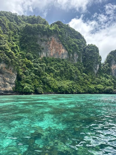 A vertical, eye-level shot captures the vibrant turquoise waters of Ao Nang, Thailand, reflecting the dramatic, verdant limestone cliffs that form the backdrop. The clear water reveals submerged rocks and corals beneath its surface. Lush green foliage blankets the towering cliffs, with patches of bare rock visible where the vegetation has not yet taken hold. The sky above is a mix of blue and white clouds. The scene appears serene and untouched, evoking a sense of natural beauty and tranquility.