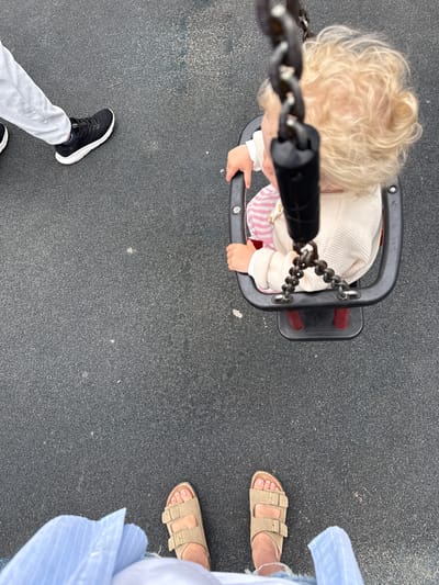 The image is an overhead shot of a child in a swing, with two adults standing on a rubber playground surface. The child, with blonde curly hair, is wearing a cream-colored knitted sweater over a pink and white striped outfit. The child's hands are resting on the black swing's safety bar. One adult is wearing black sneakers and light gray pants, with their feet visible in the lower left corner. The other adult is wearing light brown sandals and a light blue striped shirt, with their feet visible in the upper right quadrant of the image. The scene appears to be a playground, indicated by the rubber surface and swing set. The lighting suggests it is daytime. There is no visible text in the image.