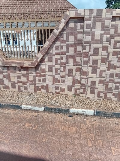 The image displays an exterior scene in Ejule, Nigeria, featuring a residential property boundary. A wall, constructed with brown and beige patterned tiles, dominates the right half of the frame. To the left, a section of a lower wall supports a metal fence, painted a light brown or gold color, featuring decorative scrollwork along the top and vertical bars below. Through the fence, a portion of a building is visible, including a white-framed window and a sloped roof covered in reddish-brown tiles. Above the wall and roofline, a segment of light blue sky with some green foliage is discernible. At the base of the tiled wall, a strip of small, light-colored gravel is present. This gravel strip is bordered by a concrete curb, painted in alternating white and black sections. The foreground consists of reddish-brown interlocking pavers, forming a pathway or road surface. No individuals or active subjects are present in the scene.