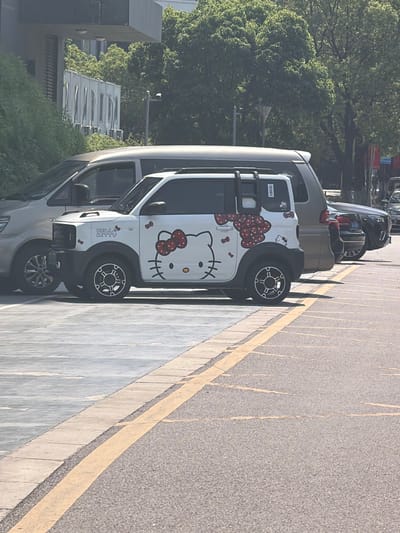 A white miniature electric vehicle adorned with large Hello Kitty graphics, including a prominent face on the side and bows on the rear quarter panel, is parked on an asphalt surface marked with yellow parking lines. The vehicle is positioned in the foreground, partially obscuring a silver minivan to its left and a dark-colored sedan to its right. The environment appears to be an outdoor parking lot, possibly in an urban setting, with a concrete sidewalk and landscaping visible in the background, suggesting it is located in the Jiangning District of China. The lighting indicates a daytime scene, likely under sunny conditions.