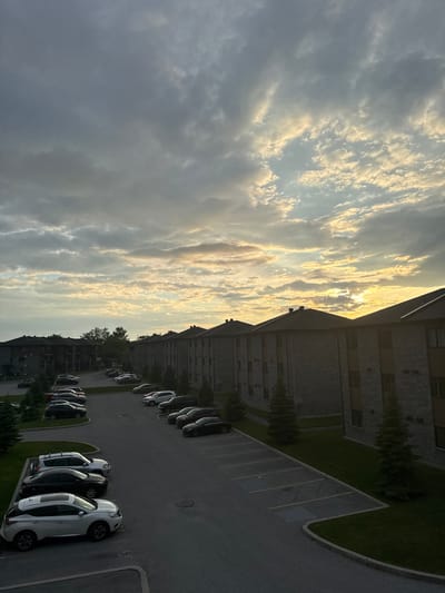 A vertical, eye-level shot captures a residential complex at dusk in Gatineau, Canada. The sky is a dramatic canvas of gray and white clouds, with patches of yellow and orange light from the setting sun peeking through. The sun is partially obscured by the clouds, creating a diffused glow that illuminates the sky.
In the foreground, a parking lot is filled with a variety of parked vehicles, including SUVs and sedans in white, black, and gray. The parking spaces are delineated by painted lines on the asphalt. Several evergreen trees are scattered throughout the parking lot and along the manicured green lawns.
The residential buildings are two to three stories tall, constructed with a mix of dark brick and wood siding. The architecture appears uniform, suggesting a modern apartment or condominium complex. Dark roofs with visible chimneys cap the buildings. The buildings are arranged in a linear fashion, with multiple units visible in the frame. Some exterior staircases are visible on the buildings. The overall scene conveys a sense of a quiet evening in a suburban or urban residential area.