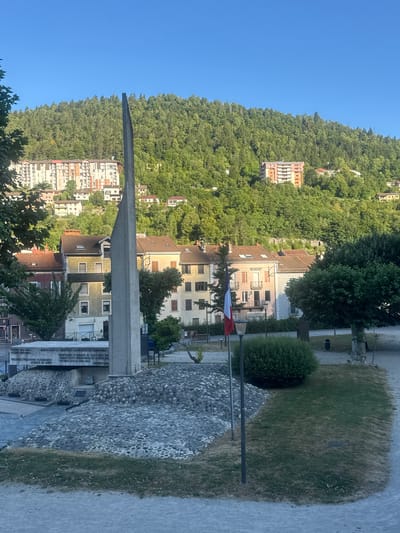 The image depicts a monument in Saint-Claude, France, with a stone obelisk and a circular base covered in cobbles. Inscriptions are visible on the base of the monument, though unreadable in detail. A French flag flies from a pole to the right of the monument. Behind the monument, residential buildings are visible, and a densely forested hill rises in the background. The sky is clear and blue. The foreground shows a path and a patch of dry grass. The scene appears to be set in a public space, likely a park or plaza.