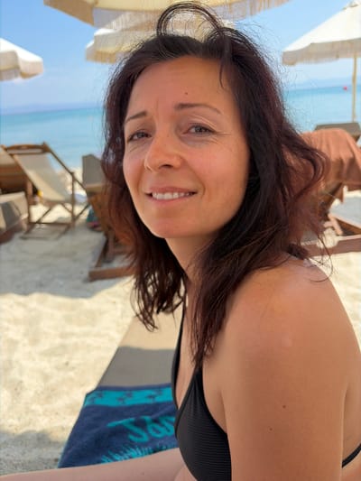 A close-up, medium shot shows a woman in a black bikini top with dark brown hair, facing forward and slightly to her left, on a sandy beach in Pallini Municipal Unit, Greece. The woman has a relaxed expression, with her mouth slightly open in a smile. The background is blurred but shows beach chairs, umbrellas, and the turquoise ocean under a clear blue sky. The lighting suggests it is a sunny day. The woman's right shoulder and upper arm are visible, with some subtle tan lines and small moles. A portion of a blue and green striped beach towel is visible in the lower left of the frame, on the sand.