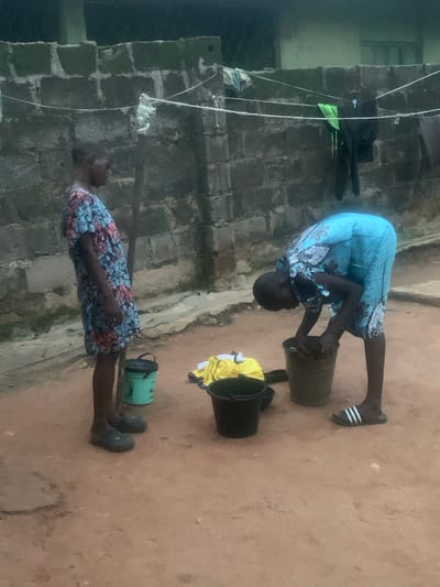 In Shagamu, Nigeria, two young individuals are engaged in laundry. A boy, facing left, stands beside a green bucket. He wears a patterned blue and pink dress and dark clogs. To his right, another individual, likely a boy, bends over a grey bucket, washing clothes with their hands. This person wears a blue patterned garment and black slides with white stripes. In the background, a brick wall is visible, with clothes hanging on a line above it. Several buckets, including a black one and a yellow garment, are on the sandy ground. The scene depicts a typical domestic chore in a semi-urban environment.