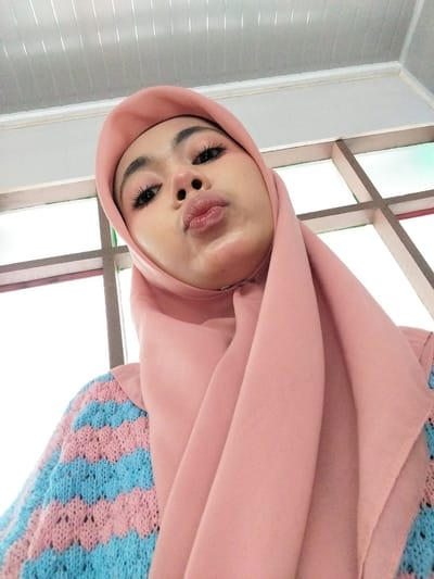 A close-up, low-angle selfie of a young woman wearing a pink hijab and a light blue and pink striped knitted sweater. Her lips are puckered, and she is looking upwards towards the camera. The background appears to be an indoor setting with a ceiling made of white, ribbed panels and a window with a grid pattern. The lighting suggests it is daytime. The location in Tibojong, Indonesia, is not explicitly visible in the image.