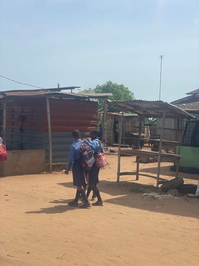 The image shows a dirt road in Ikoga, Nigeria. Two children in school uniforms are walking along the road, each carrying a backpack. Buildings are present on either side of the road, constructed from brick, corrugated metal, and wood. An antenna is seen extending from the roof of one of the buildings.