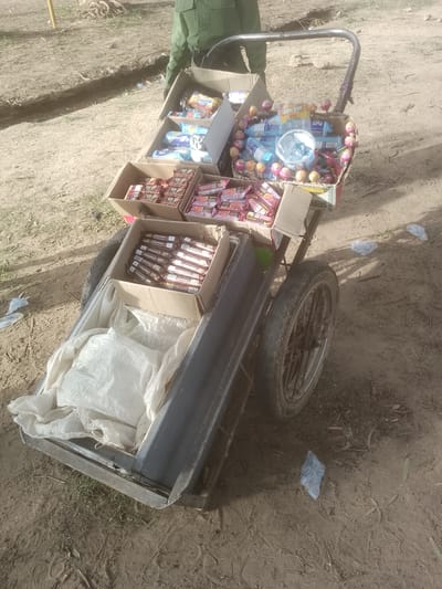 In Majemu, Nigeria, a mobile vending cart is laden with an assortment of confectionery items. The cart, a two-wheeled affair, is positioned on a dirt path, suggesting an outdoor market or roadside setting. The items for sale are primarily packaged snacks, including chocolate bars, biscuits, and candies, some of which are individually wrapped lollipops. These are displayed in cardboard boxes and plastic containers on the cart. The cart itself appears to be constructed from metal, with a handle for pushing and two wheels. A person in a green uniform, possibly a vendor or a uniformed worker, is partially visible in the upper background, standing behind the cart. The surrounding environment is dusty and dry, with sparse vegetation and some scattered debris, including plastic bags, visible on the ground. The overall impression is one of a typical street-side commerce scene in a rural or semi-urban Nigerian location.