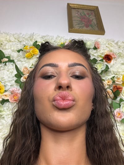 A young woman with dark, wavy hair and full lips puckered for a kiss poses for a selfie in front of a floral wall. She is wearing shimmery eyeshadow and has blush on her cheeks. The floral wall is composed of white, yellow, pink, and orange artificial flowers. A small, framed picture of red flowers hangs on the wall behind her. The scene is indoors, and the lighting appears to be artificial. The location is unspecified within the city of Novi Iskar, Bulgaria.