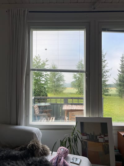 The image shows an interior view through a window onto a balcony and the surrounding landscape. In the foreground, a plush armchair covered with a white textured blanket and a dark furry throw is visible on the left. A purple octopus plush toy with a single eye and a tag that reads "KONG" rests on the blanket. Next to it is a potted plant with long, green leaves and a small, square reflective object. A large framed artwork leaning against the wall depicts a stylized landscape with white-roofed buildings, trees, and abstract fields in earthy tones.

The window, divided into two main sections, reveals a balcony with wooden flooring and a dark railing. On the balcony, there is a round glass-topped table with two chairs and a rectangular wooden table with a bench. Beyond the balcony, a green lawn stretches out, dotted with trees. In the distance, a field of yellow crops is visible, bordered by more trees and a hazy, overcast sky. Two tall evergreen trees are prominent on the right side of the frame.

The overall scene suggests a peaceful, perhaps rural, setting, potentially a residential area in Hämeenlinna, Finland, given the architectural style of the buildings in the artwork and the northern European landscape features. The light is diffused, indicating an overcast or early morning/late afternoon time of day. There are no people or animals actively engaged in any actions within the frame.